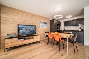 A modern living room with a wooden wall, a flat-screen TV, a wooden dining table with orange chairs, and a kitchen area in the background.