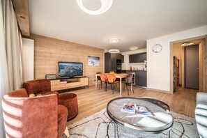 A modern living room with a flat-screen TV, a dining area with orange chairs, and a kitchen in the background.