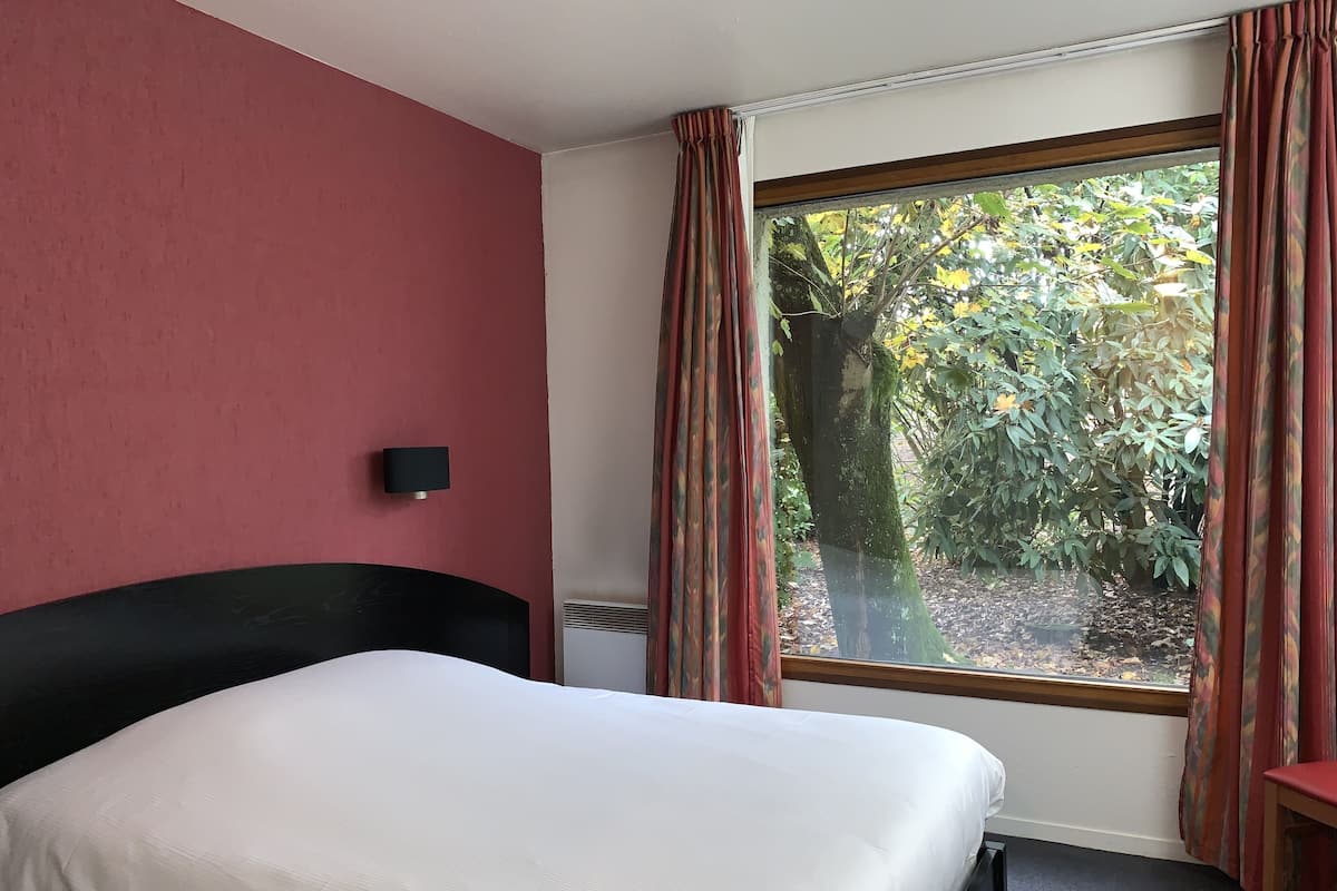 A hotel room with a bed, a window with curtains, a wall-mounted light, and a view of trees outside.