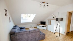 A modern living room with a grey sofa, a small white table, a TV on a white stand, and a floor lamp. - Modern and bright vacation apartment with Hutewald view (Vöhl)