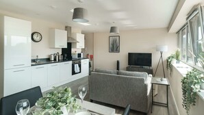 Uma sala de estar moderna com um sofá cinza, uma mesa de jantar com cadeiras, uma televisão e uma área de cozinha com armários e eletrodomésticos.