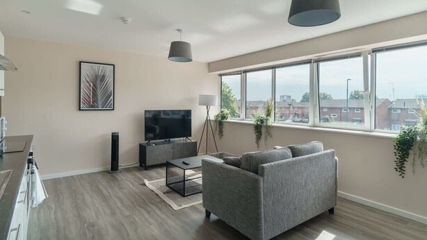 Uma sala de estar moderna com um sofá cinzento, uma televisão de tela plana, uma mesa de centro e uma grande janela com vista para edifícios.