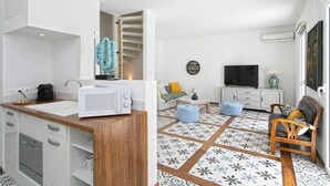 Een moderne keuken met een magnetron, spoelbak en kastjes, een zithoek met een bank en televisie, en vloerbedekking met een patroon.