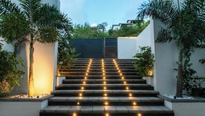 Eine Treppe mit gleichmäßig angeordneten Lichtern, die zu einem begrenzten Eingang führt, umgeben von Pflanzen und einer weißen Mauer.