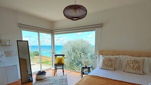 Un dormitorio con cama, una silla, una mesita, un ventanal con vistas al océano y un ventilador de techo.
