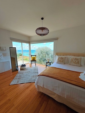 A bedroom with a bed, a chair, a small table, a large window with a view of the ocean, and a ceiling fan.