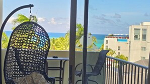 Un balcon avec un fauteuil suspendu, une table et une vue sur l’océan et les bâtiments.