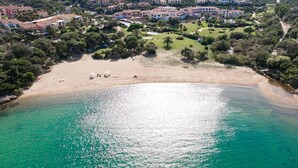 An aerial view of a coastal area with a beach, clear turquoise waters, and a residential complex with multiple buildings. - Shared Pool Apartment in Pula (Pula (CA))