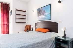 A bedroom with a bed, a nightstand, a wall-mounted shelf, a framed picture, and a red curtain.