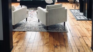 Un salón con un sofá verde oscuro, dos sillones blancos, una araña, ventanas amplias con cortinas blancas y suelo de madera.