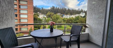 A balcony with a round table and two chairs, a potted plant, and a view of buildings and trees.