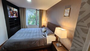 Quarto de hotel com uma cama grande, uma televisão na parede esquerda, uma janela com cortinas, uma mesinha de cabeceira com abajur e uma parede com padrão geométrico.