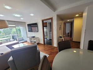 Uma sala de estar moderna com sofá, mesa de jantar e televisão. Possui uma varanda com área de estar ao ar livre e vista para a cidade.