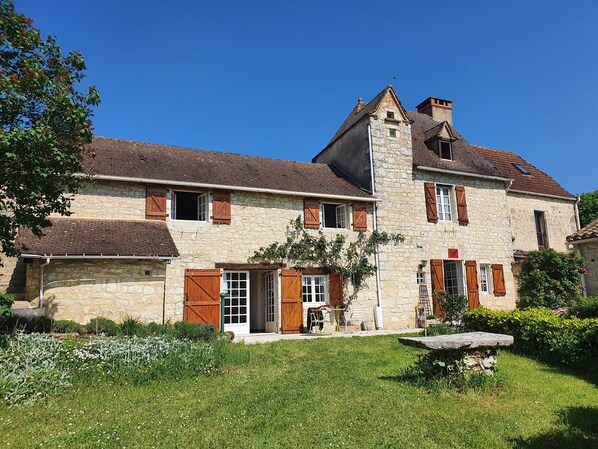 Une maison en pierre avec une grande porte d’entrée, des volets en bois et une cheminée en pierre.