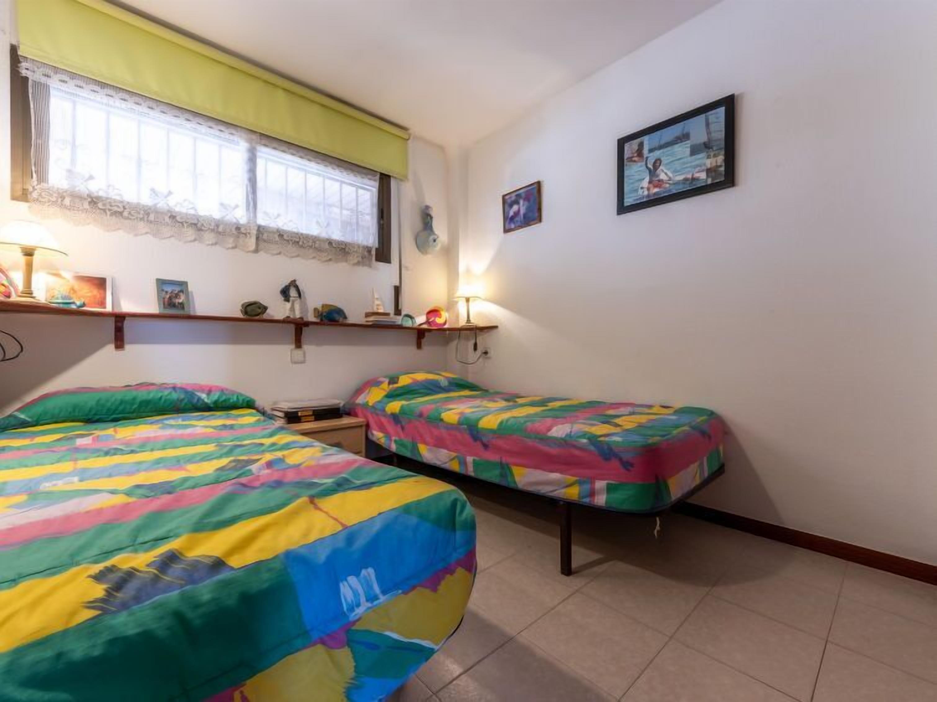 A room with two beds, a window with a curtain, a shelf with various items, and a wall with two framed pictures.