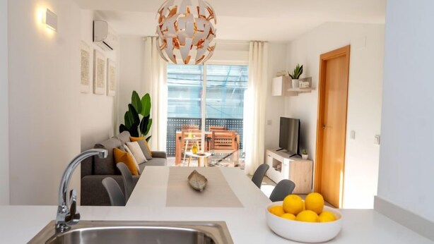 A modern kitchen with a large sink, a dining area with a table and chairs, a sofa with cushions, a television, and a plant. - Apartment Sakura by Interhome (Segur de Calafell)