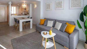 A modern living room with a grey sofa, a small white side table, and a dining area with a white table and chairs. - Apartment Sakura by Interhome (Segur de Calafell)