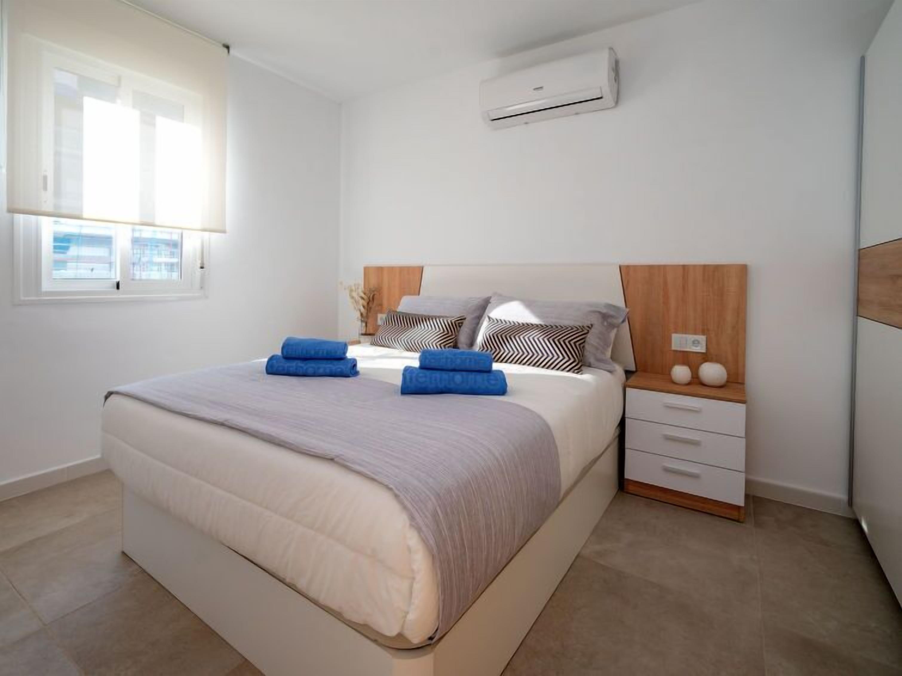 Une chambre moderne avec un grand lit, une table de chevet, un climatiseur fixé au mur et une fenêtre avec des stores.