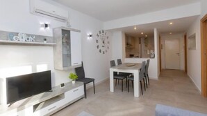 A modern living room with a flat-screen TV, a dining table with chairs, a kitchen visible through an open door, and a clock on the wall. - Apartment Karma by Interhome (Segur de Calafell)