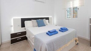 A modern hotel room with a bed, bedside tables, a nightstand, a window with blinds, and an air conditioning unit. - Apartment Karma by Interhome (Segur de Calafell)