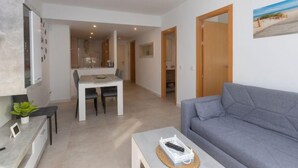 A modern living room with a grey sofa, a dining table with chairs, a kitchen area, and a bathroom visible through an open door. - Apartment Karma by Interhome (Segur de Calafell)