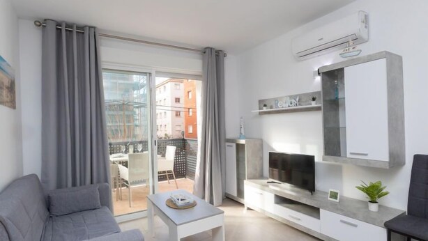 A modern living room with a grey sofa, a small white table, a TV on a white cabinet, and a balcony with grey curtains. - Apartment Karma by Interhome (Segur de Calafell)