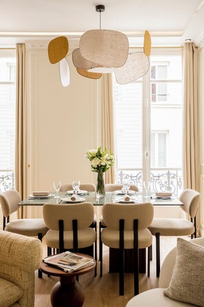 Un coin repas avec une table ronde dressée pour six personnes, entourée de six chaises. On peut voir un vase rempli de fleurs sur la table, ainsi qu’un magazine sur une petite table d’appoint.