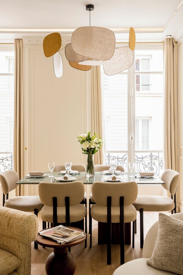 A dining area with a round table set for six, surrounded by six chairs. There is a vase with flowers on the table, and a magazine on a side table.