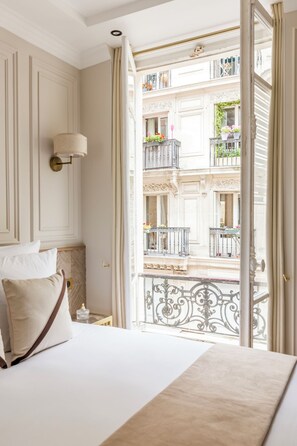 A hotel room with a bed, a nightstand, and a view of a balcony with ornate railings and potted plants.