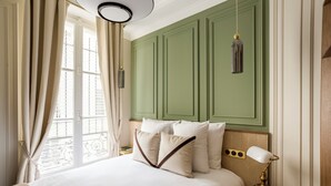 A hotel room with a large bed, green paneling on the walls, a window with curtains, and a bedside table with a lamp and a small tray. - Merveil - Luxury Suite - Marais - Caire I (Paris)