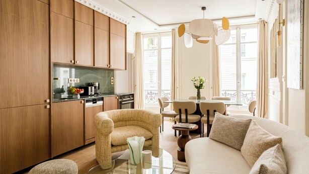 A modern living room with a sofa, armchairs, a glass coffee table, and a kitchen area with wooden cabinets and appliances. - Merveil - Luxury Suite - Marais - Caire I (Paris)
