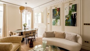 A well-lit living room with a sofa, chairs, a glass coffee table, and a dining area with a table and chairs. There are large abstract paintings on the walls. - Merveil - Luxury Suite - Marais - Caire I (Paris)