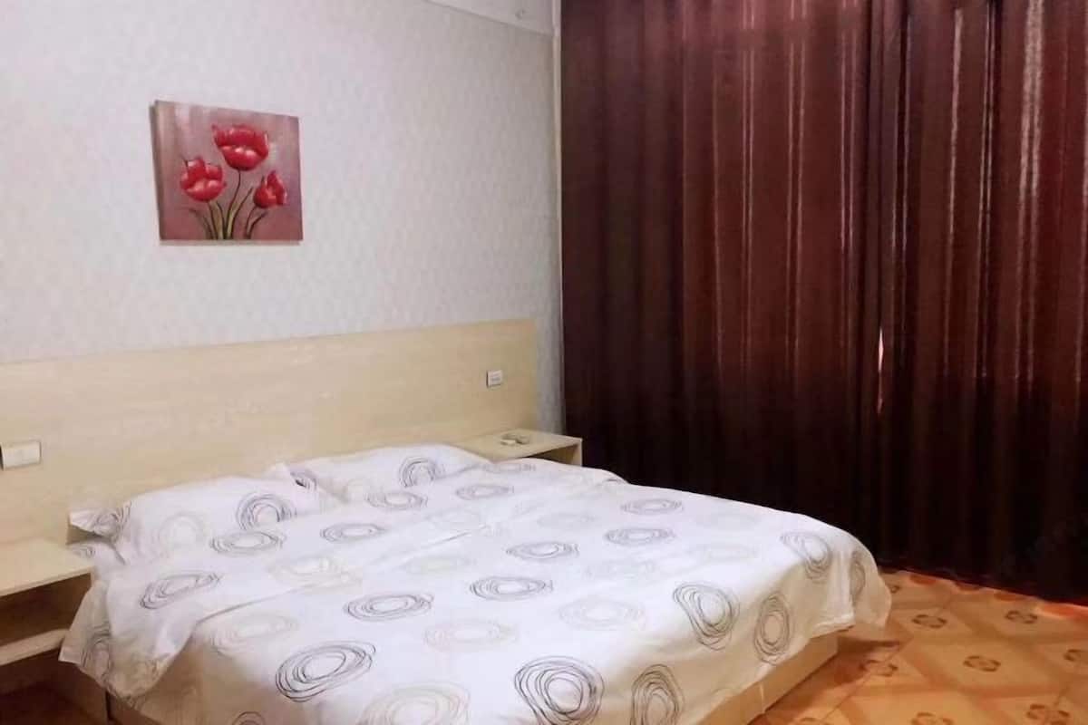 A bedroom with a bed, a nightstand, a wall-mounted telephone, and a window with curtains.