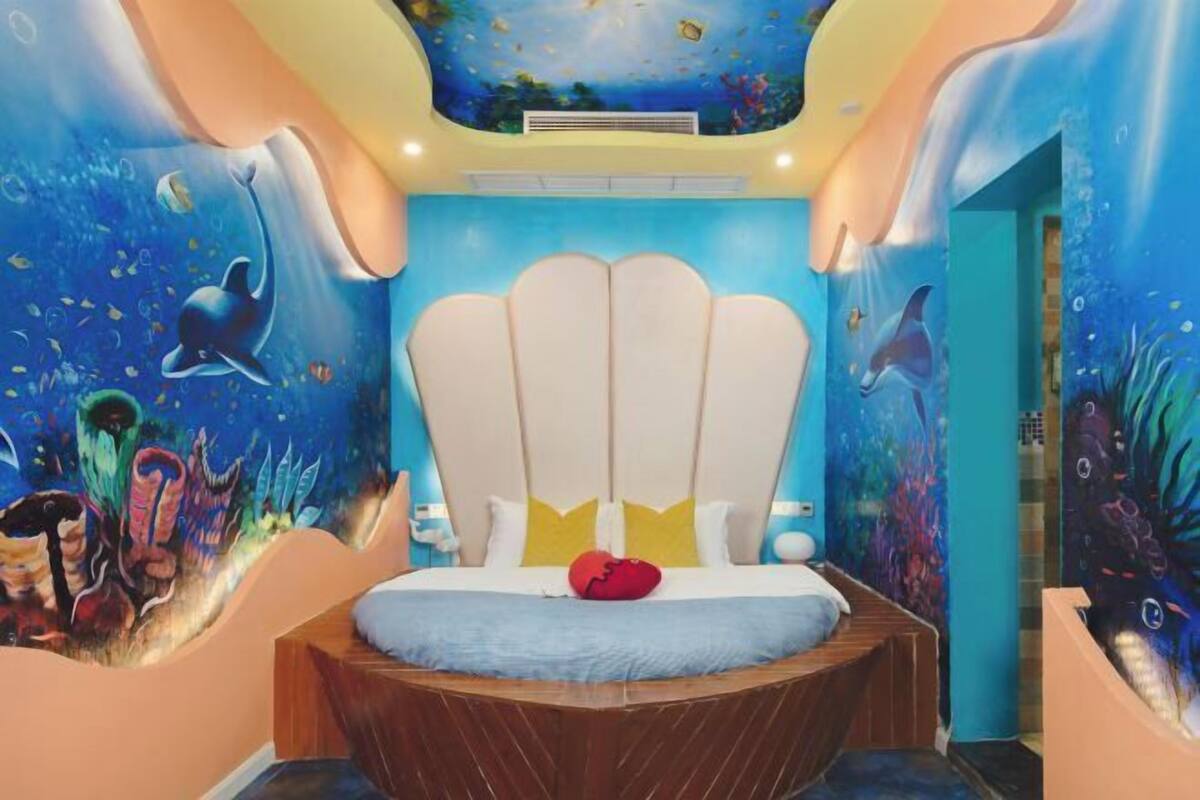 A bedroom with a bed centered under a large, scalloped headboard, surrounded by vibrant underwater-themed murals.