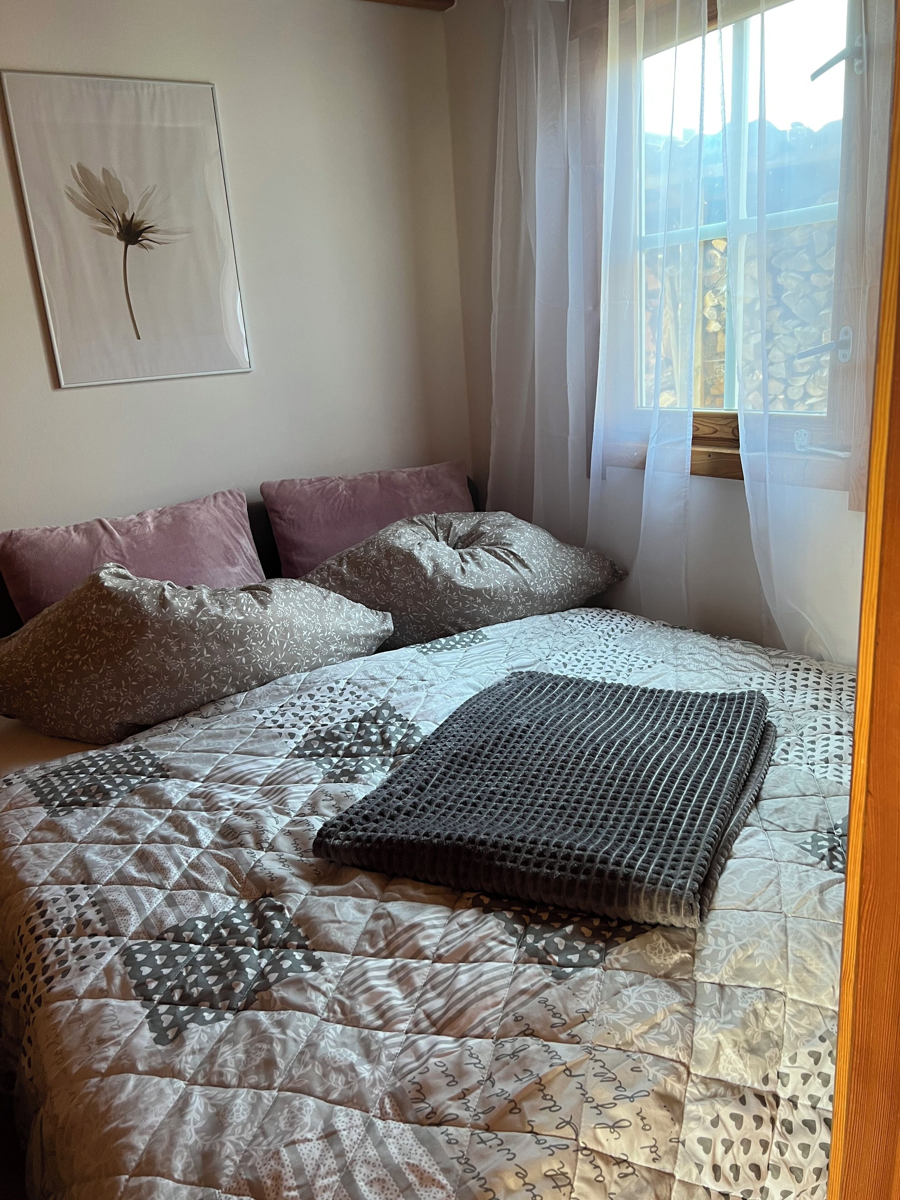 A neatly made bed with patterned bedding and pillows, a framed artwork on the wall, and a window with sheer curtains.