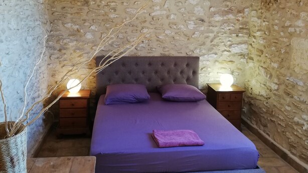 Camera da letto con parete in pietra, trave in legno a soffitto, testiera imbottita, letto con biancheria viola, due comodini con lampade e un cesto in vimini.