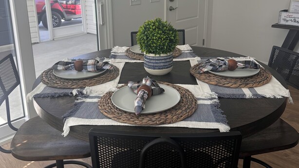 Un coin repas avec une table ronde, des chaises noires, des sets de table en fibres tressées et une plante en pot.