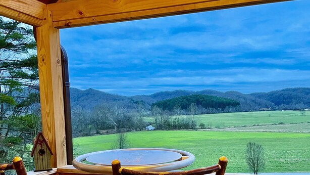 A wooden deck with a round table and two chairs, overlooking a green landscape with hills.