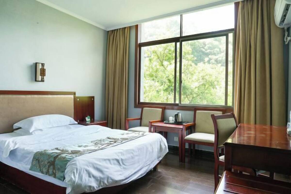 A hotel room with a bed, a small table, two chairs, a window with curtains, and a wall-mounted light fixture.