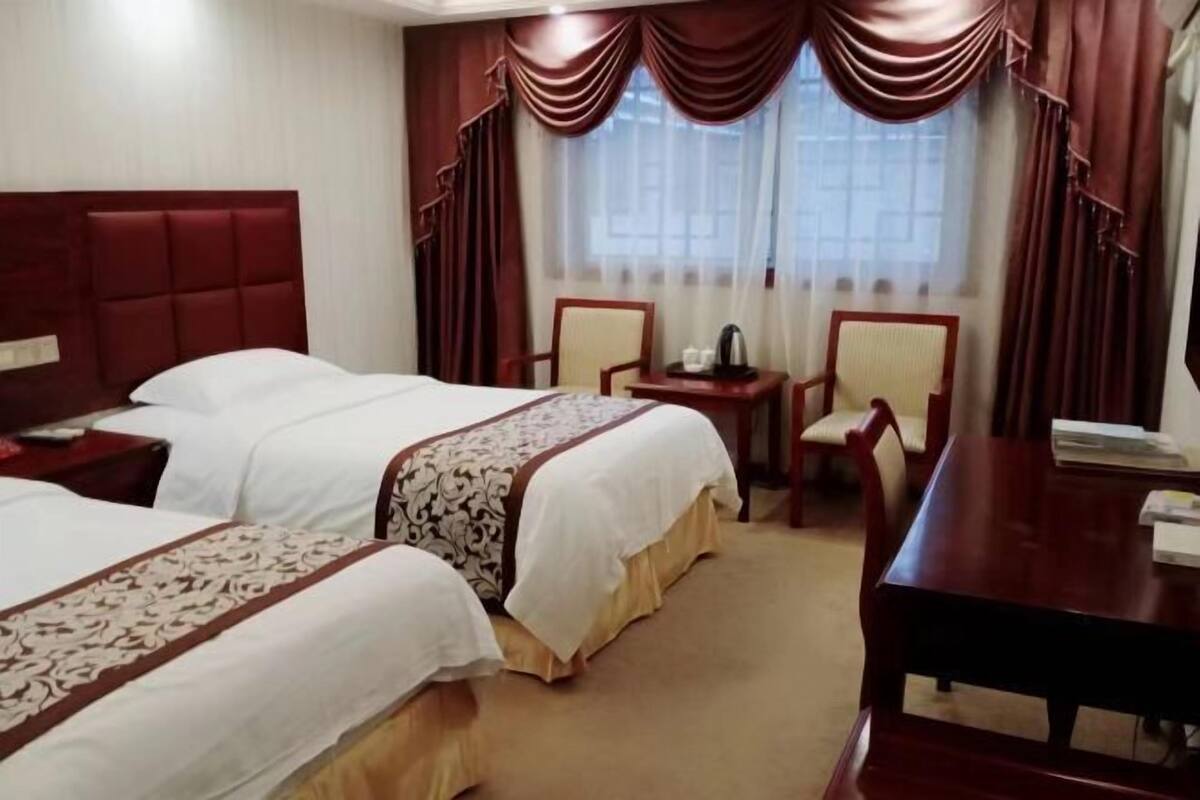 A hotel room with two beds, a desk, a chair, and a small table with a kettle.