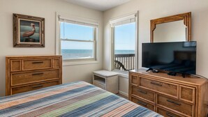 A bedroom with a bed, dresser, TV, and a window with a view of the ocean. - Golden Strand Unit J2: Oceanfront | Shared Pool (Kill Devil Hills)