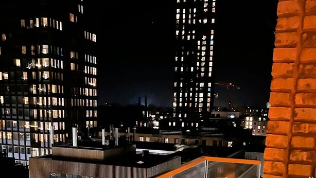 A nighttime view of a modern building with multiple illuminated windows, a glass railing, and a brick wall with a potted plant. - City Apartment in Copenhagen with 1 bedrooms sleeps 2 (Copenhagen)