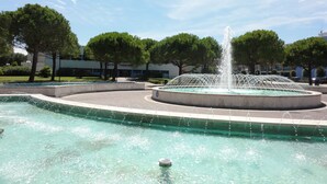 Une fontaine avec des jets d’eau dans un bassin circulaire, entourée d’arbres et de bâtiments.