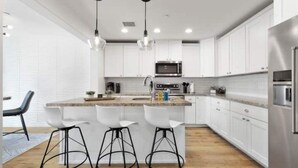 Une cuisine moderne avec des armoires blanches, un réfrigérateur en acier inoxydable, un four à micro-ondes et un comptoir en bois. On y trouve trois tabourets de bar et des luminaires suspendus au-dessus de l’îlot central.