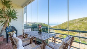 A balcony with a wooden table and chairs, a potted plant, and a view of a hilly landscape.