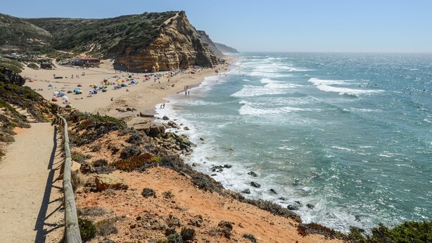 A coastal beach with a dirt path leading to the shore, a cliff, and a building in the background.