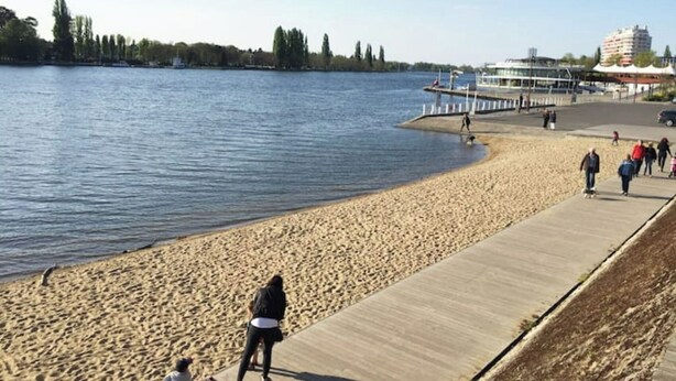 Ein sandiger Strand mit einer Betonpromenade, einem Steg und Gebäuden im Hintergrund.