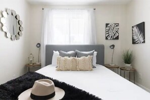 Une chambre à coucher avec un lit, un tapis noir à poils longs, un chapeau, un miroir, une fenêtre avec des rideaux et des œuvres d’art murales.