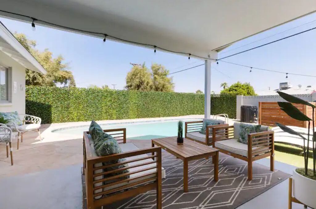 A covered outdoor patio with a pool, wooden furniture, and a hedge in the background.