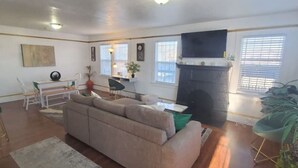 A living room with a beige sofa, a dining table with chairs, a fireplace, and large windows with blinds. - The Modern Family Getaway (Oakland)